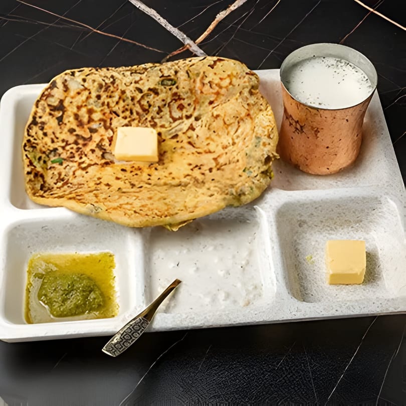 Indian paratha with butter, green chutney, white sauce, and a copper cup of lassi on a white tray.