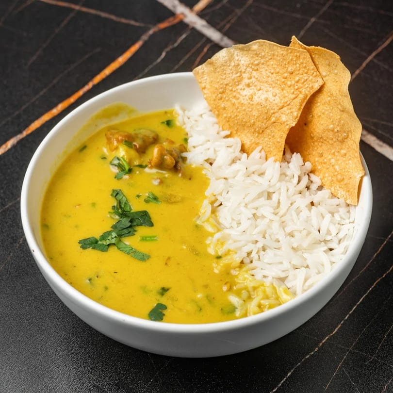 A white bowl of Lassi Lounge's yellow Indian curry, white rice, and two papadums.